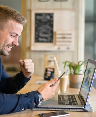 A man celebrating as he reads good news on his phone beside his laptop